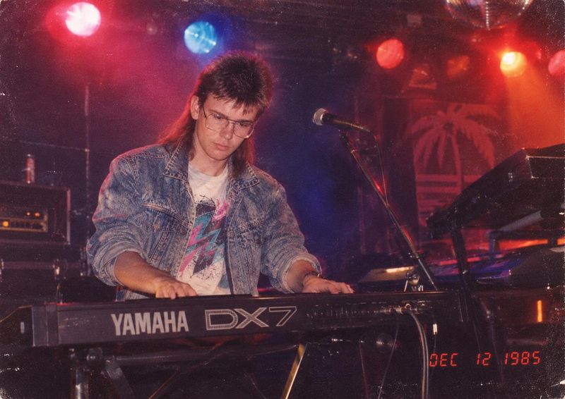 Tommy Cape on a small stage, head bowed over a Yamaha DX7 synthesizer, in an acid-wash denim jacket under red and blue stage lights.