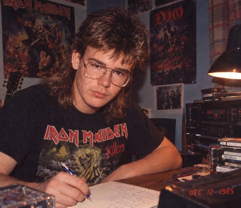 Tommy Cape at his desk writing lyrics in a spiral notebook and glancing up at the camera, with heavy-metal posters on the wall, a desk lamp, a cassette deck and a stack of tapes beside him.
